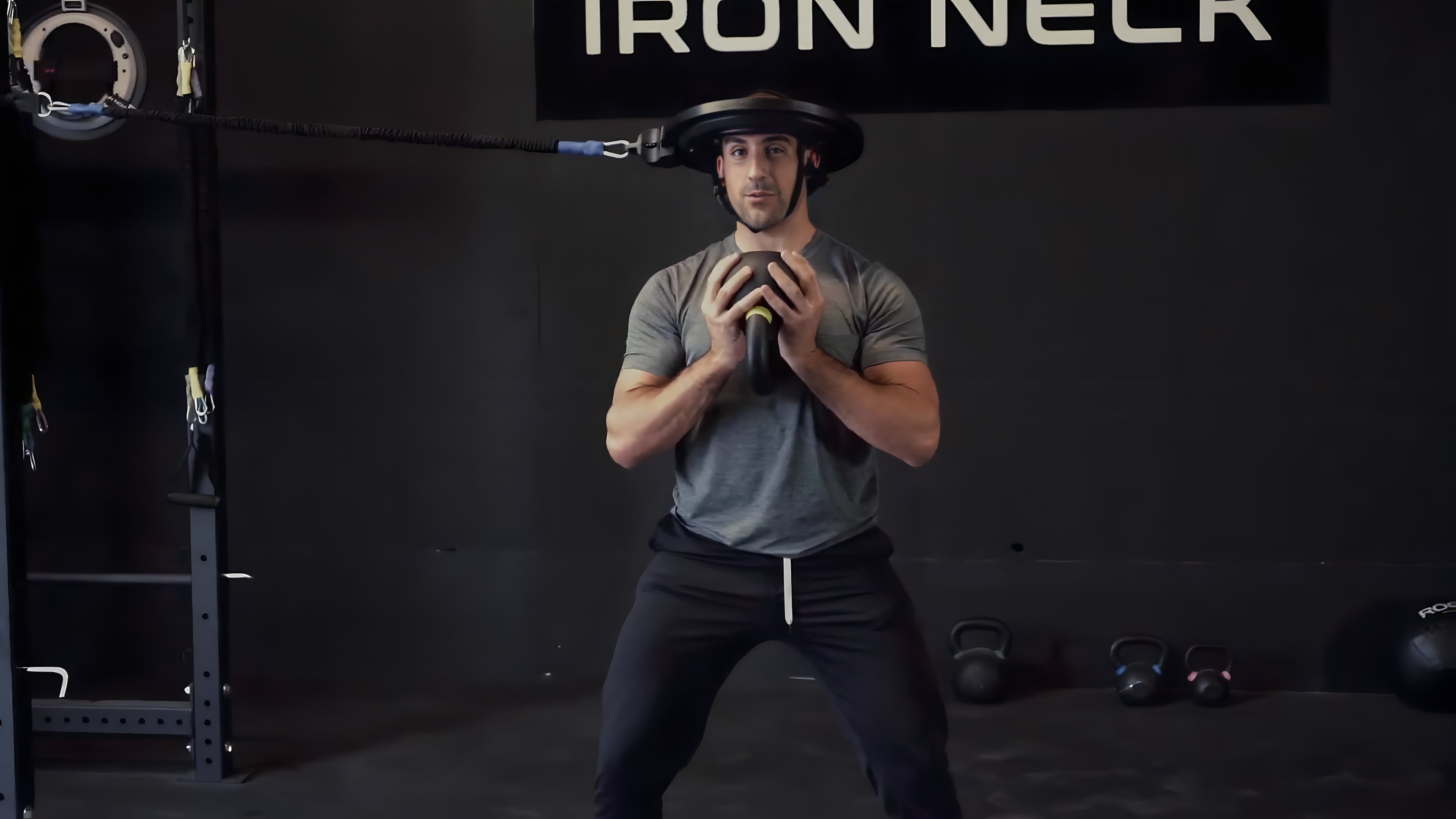 Mike Salemi wears Iron Neck on his head and holds a kettlebell with both hands on his chest.